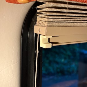 May include: Close-up of a window with a beige pleated shade and a black frame. The shade is partially drawn, revealing a glimpse of the outside. An orange fabric panel is visible above the window.