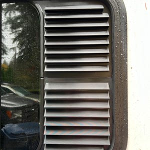 May include: A black plastic vent with horizontal slats, mounted on a white surface. The vent is closed.