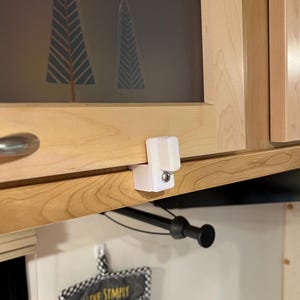 May include: White plastic cabinet latch with a silver knob. The latch is mounted on a wooden cabinet door.