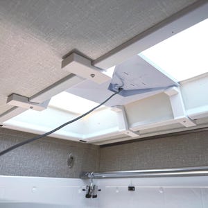May include: A white plastic skylight shade mount installed on a textured grey ceiling surrounding a rectangular skylight. The assembly features white brackets, a central white panel with a thin black wire, and a metal curtain rod below.
