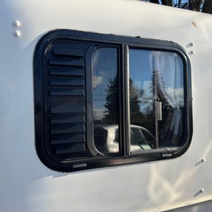 May include: A rectangular window with a black frame and a black louvered sunshade is set into the white exterior of a vehicle. The window glass reflects the surrounding environment, including trees and a car.