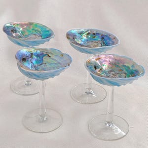 May include: Set of four iridescent abalone shell bowls on clear glass stems. The bowls are a light blue colour with iridescent green, blue, and purple hues.
