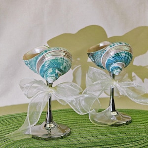 May include: Two decorative wedding toasting glasses made from seashells with a teal and white pattern. The glasses are on a green surface and have silver metal stems.