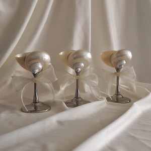 Könnte beinhalten: Drei weiße Muschel-Kerzenhalter mit silbernen Ständern und durchsichtigen weißen Schleifen.
