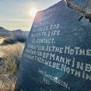 May include: A gray rock with white painted text that reads "THE ROCH OF FAIHT AND TRUNT NATURE IS.COD THE YEY. TO. LIFE. IS. CONTACT. EVOLUTION IS THE MOTHER AND FATHER OF MANKIND. WITHOUT THEM.WE.BE NOTHIN JOHN. SAMUELSON 1927."