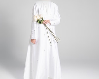 May include: A white long-sleeved dress with a V-neckline and a subtle pattern of small white dots. The dress has a flowing skirt and is worn with white sandals.