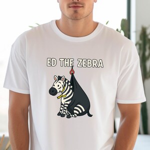 May include: White t-shirt featuring a cartoon zebra named Ed. The zebra has black and white stripes, a yellow collar, and is hanging from a black sack. The text "ED THE ZEBRA" is above the zebra.