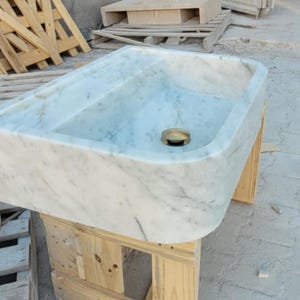 May include: Rectangular marble sink with a light gray and white veined pattern. The sink has a rounded front edge and a brass drain. It is sitting on a wooden pallet.