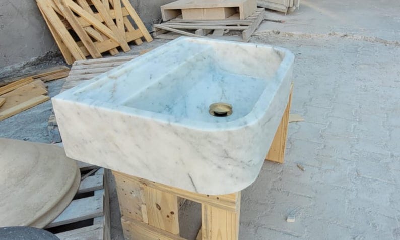 May include: Rectangular marble sink with a light gray and white veined pattern. The sink has a rounded front edge and a brass drain. It is sitting on a wooden pallet.