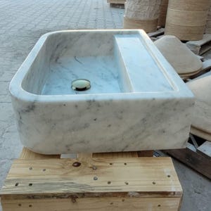 May include: A rectangular marble sink with rounded corners and a built-in shelf. The sink is white with gray veining and a gold-colored drain. It sits on a wooden pallet, with other stone elements in the background.