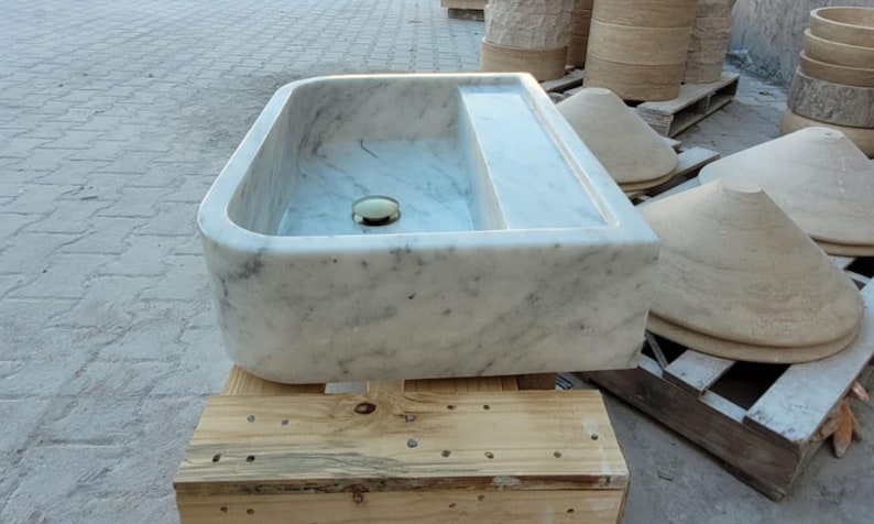 May include: A rectangular marble sink with rounded corners and a built-in shelf. The sink is white with gray veining and a gold-colored drain. It sits on a wooden pallet, with other stone elements in the background.