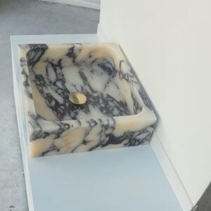 Puede incluir: Un lavabo cuadrado de m&aacute;rmol con un desag&uuml;e dorado. El lavabo presenta un patr&oacute;n veteado crema y gris. El lavabo est&aacute; empotrado en una superficie azul claro y est&aacute; junto a una pared blanca.