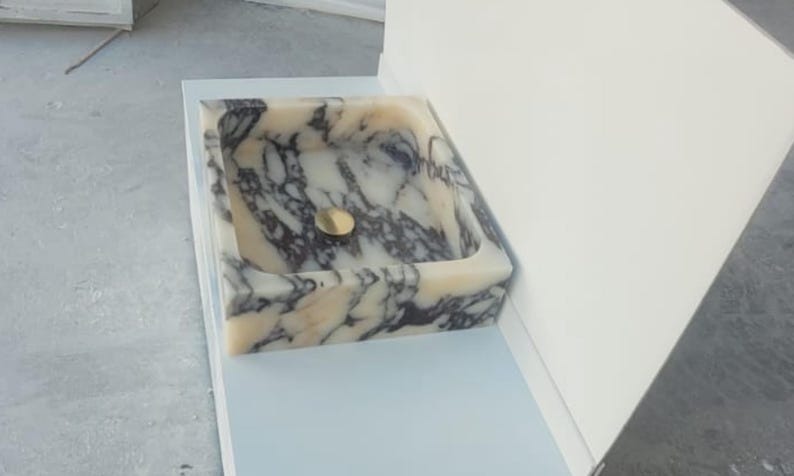 Puede incluir: Un lavabo cuadrado de m&aacute;rmol con un desag&uuml;e dorado. El lavabo presenta un patr&oacute;n veteado crema y gris. El lavabo est&aacute; empotrado en una superficie azul claro y est&aacute; junto a una pared blanca.