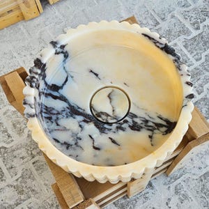 May include: A white marble bowl with black and brown veining. The bowl has a scalloped edge and a small hole in the centre.
