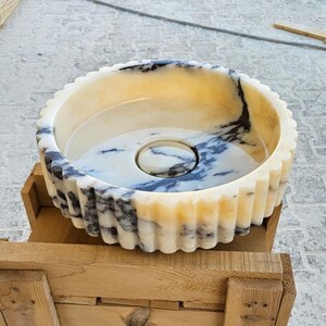 May include: A white marble sink with black veining, featuring a fluted rim and a circular drain. The sink is sitting on a wooden crate.