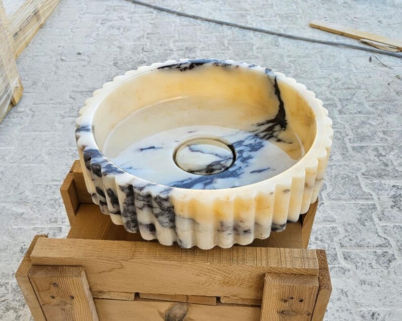 May include: A white marble sink with black veining, featuring a fluted rim and a circular drain. The sink is sitting on a wooden crate.