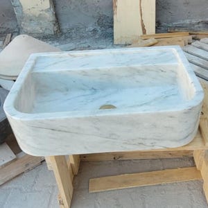 May include: A rectangular marble sink with a divided basin, featuring a light gray and white veined pattern. The sink is set on a wooden stand, showcasing a natural stone design. Other stone objects are visible in the background.