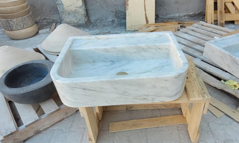 May include: A rectangular marble sink with a divided basin, featuring a light gray and white veined pattern. The sink is set on a wooden stand, showcasing a natural stone design. Other stone objects are visible in the background.