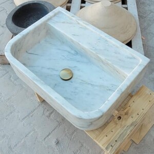 May include: A rectangular marble sink with a brass drain, showcasing a light gray and white veined pattern. The sink is set on a wooden pallet, with other stone basins visible in the background. A modern bathroom fixture.