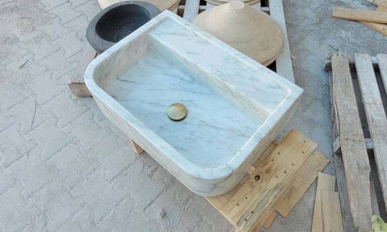 May include: A rectangular marble sink with a brass drain, showcasing a light gray and white veined pattern. The sink is set on a wooden pallet, with other stone basins visible in the background. A modern bathroom fixture.