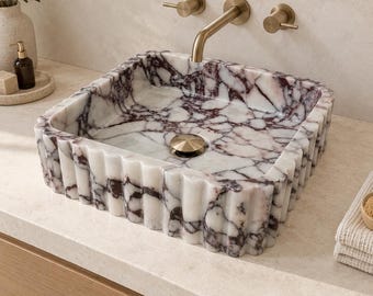 May include: A square bathroom sink crafted from white marble with prominent dark purple veining. The exterior features a vertical fluted texture, and the basin includes a centred metallic drain cover in a brushed brass finish.