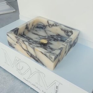 Puede incluir: Un lavabo rectangular de m&aacute;rmol con un desag&uuml;e dorado. El lavabo tiene un patr&oacute;n veteado beige y gris. El lavabo se exhibe sobre un soporte blanco con el texto "NOM NATURAL MARBLE" grabado en la parte delantera.