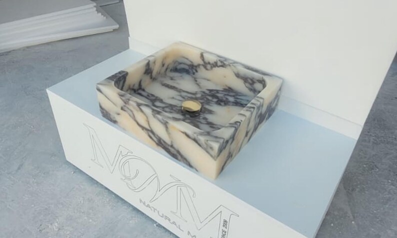 Puede incluir: Un lavabo rectangular de m&aacute;rmol con un desag&uuml;e dorado. El lavabo tiene un patr&oacute;n veteado beige y gris. El lavabo se exhibe sobre un soporte blanco con el texto "NOM NATURAL MARBLE" grabado en la parte delantera.