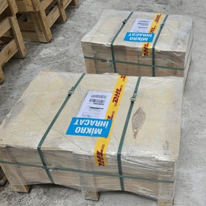 May include: Two wooden crates wrapped in clear plastic and secured with green straps. Each crate has a DHL shipping label and a blue sticker with white text. The crates are on a concrete surface.