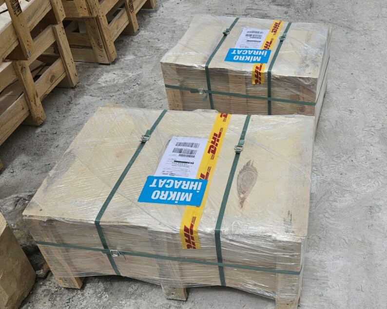 May include: Two wooden crates wrapped in clear plastic and secured with green straps. Each crate has a DHL shipping label and a blue sticker with white text. The crates are on a concrete surface.