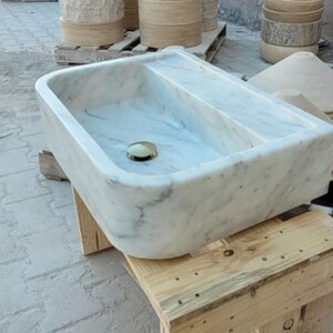 May include: A rectangular marble sink with a divided basin and a brass drain. The sink is white with gray veining and sits on a wooden pallet. Other stone objects are in the background.