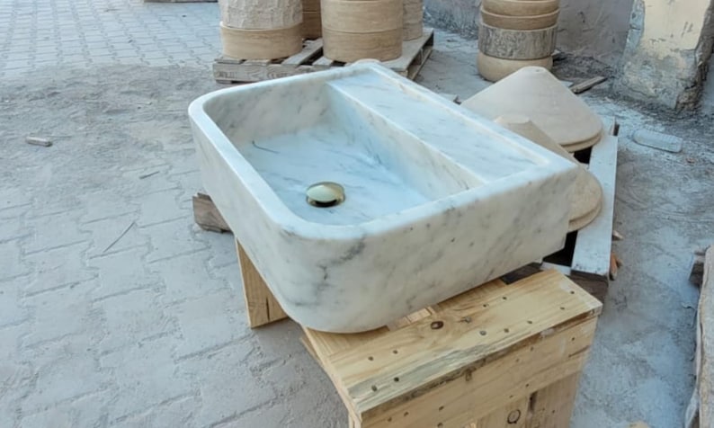 May include: A rectangular marble sink with a divided basin and a brass drain. The sink is white with gray veining and sits on a wooden pallet. Other stone objects are in the background.