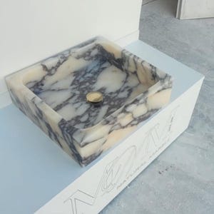 Puede incluir: Lavabo rectangular de m&aacute;rmol con desag&uuml;e dorado. El lavabo presenta un patr&oacute;n veteado crema y gris. Est&aacute; colocado sobre una superficie blanca con el texto "NATURAL MARBLE" visible. El lavabo es un accesorio de ba&ntilde;o.