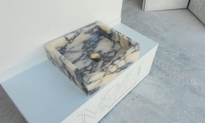 Puede incluir: Lavabo rectangular de m&aacute;rmol con desag&uuml;e dorado. El lavabo presenta un patr&oacute;n veteado crema y gris. Est&aacute; colocado sobre una superficie blanca con el texto "NATURAL MARBLE" visible. El lavabo es un accesorio de ba&ntilde;o.