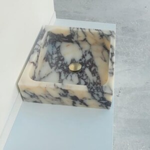 Puede incluir: Un lavabo rectangular de m&aacute;rmol con un desag&uuml;e dorado. El lavabo tiene una base de color crema con vetas grises oscuras y negras. El lavabo est&aacute; empotrado en una superficie blanca.