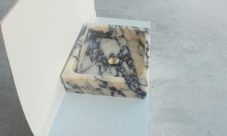 Puede incluir: Un lavabo rectangular de m&aacute;rmol con un desag&uuml;e dorado. El lavabo tiene una base de color crema con vetas grises oscuras y negras. El lavabo est&aacute; empotrado en una superficie blanca.