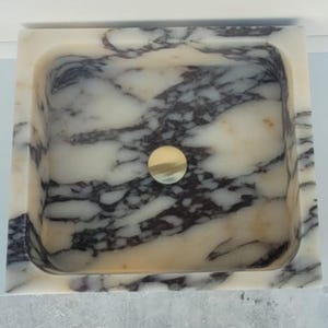 Puede incluir: Un lavabo cuadrado de m&aacute;rmol con un desag&uuml;e dorado. El lavabo presenta un patr&oacute;n veteado blanco y gris. El lavabo est&aacute; colocado contra una pared azul claro.