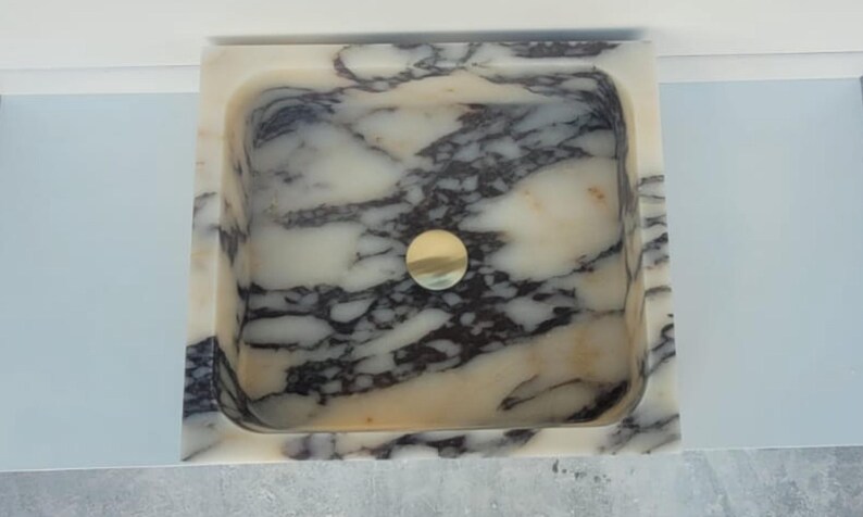 Puede incluir: Un lavabo cuadrado de m&aacute;rmol con un desag&uuml;e dorado. El lavabo presenta un patr&oacute;n veteado blanco y gris. El lavabo est&aacute; colocado contra una pared azul claro.