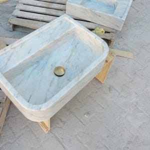 May include: A rectangular marble sink with a brass drain. The sink has a light gray and white veined pattern. Other stone basins and wooden pallets are in the background. The sink is supported by a wooden stand.