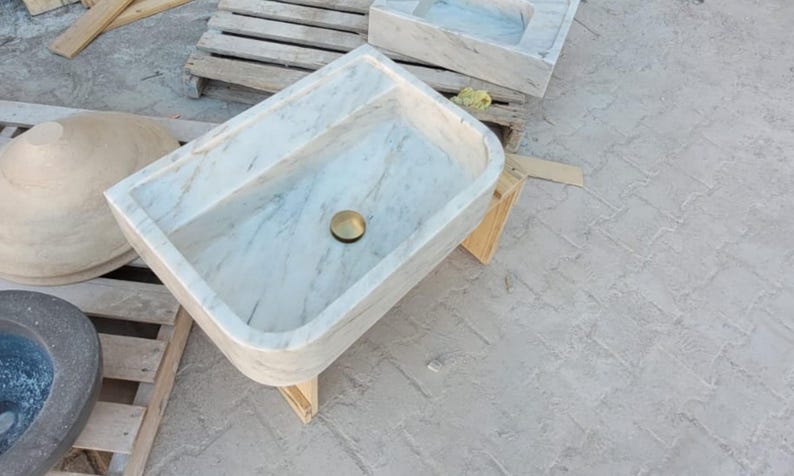May include: A rectangular marble sink with a brass drain. The sink has a light gray and white veined pattern. Other stone basins and wooden pallets are in the background. The sink is supported by a wooden stand.