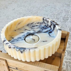 May include: A white marble sink with black veining. The sink has a fluted edge and a round drain.