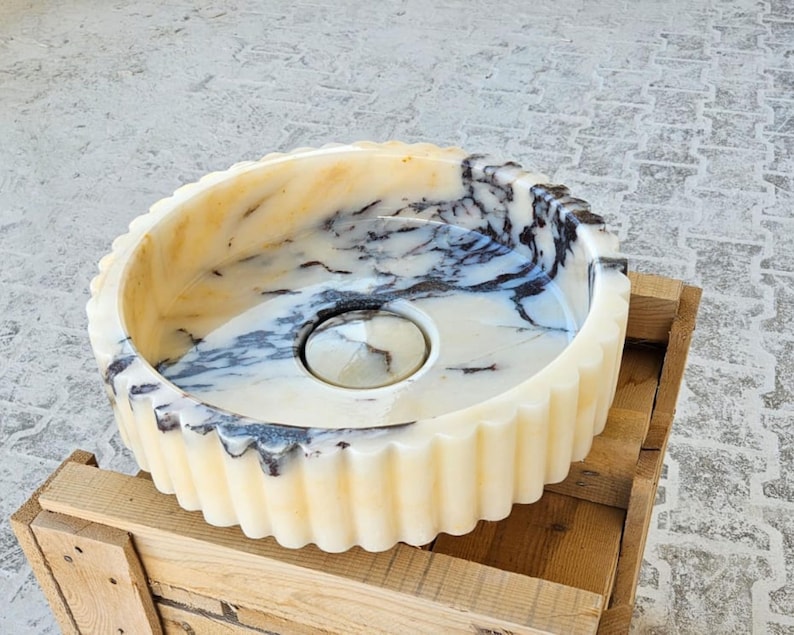May include: A white marble sink with black veining. The sink has a fluted edge and a round drain.