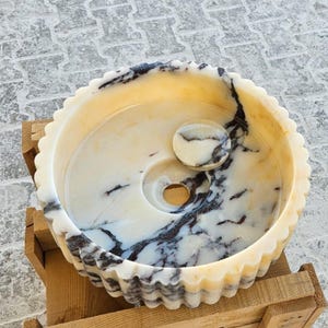 May include: A white marble sink with black veining, featuring a fluted rim and a small round marble drain stopper. The sink is set on a wooden base.