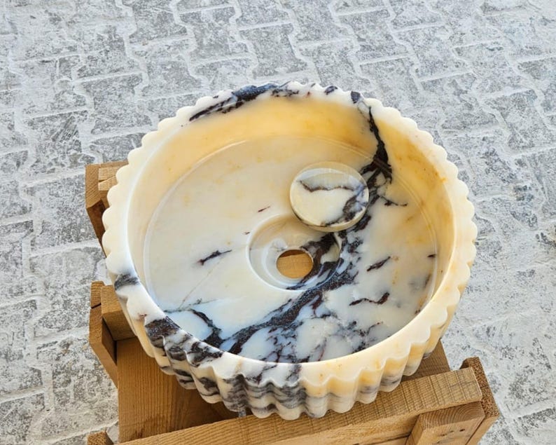 May include: A white marble sink with black veining, featuring a fluted rim and a small round marble drain stopper. The sink is set on a wooden base.