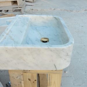 May include: A rectangular marble sink with a rounded front edge. The sink has a light gray and white veined pattern and a gold-colored drain. It is set on a wooden pallet.