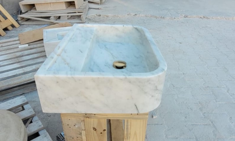 May include: A rectangular marble sink with a rounded front edge. The sink has a light gray and white veined pattern and a gold-colored drain. It is set on a wooden pallet.