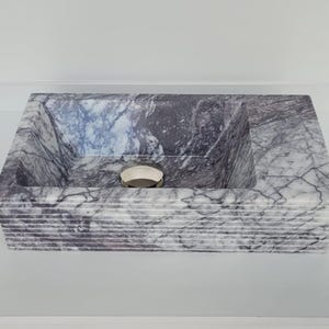 Puede incluir: Un lavabo rectangular de mármol blanco con un patrón de vetas grises y moradas. El lavabo tiene un borde texturizado en capas y un desagüe redondo de plata.