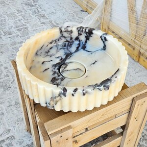 May include: A white marble sink with black veining, featuring a fluted rim and a round shape. The sink is sitting on a wooden pallet.