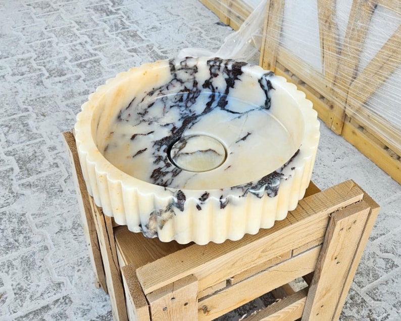 May include: A white marble sink with black veining, featuring a fluted rim and a round shape. The sink is sitting on a wooden pallet.