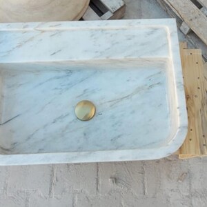 May include: Rectangular marble sink with a light gray and white veined pattern. The sink has a built-in shelf and a gold-colored drain. The sink is on a concrete surface.