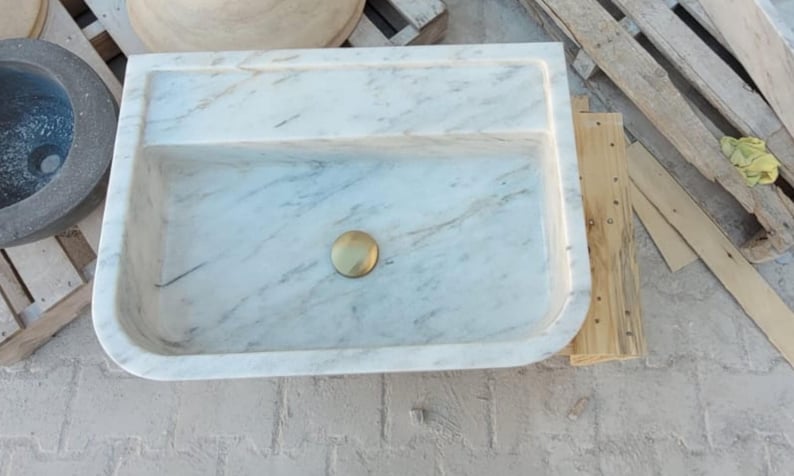May include: Rectangular marble sink with a light gray and white veined pattern. The sink has a built-in shelf and a gold-colored drain. The sink is on a concrete surface.